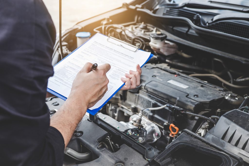 A technician with a clipboard going over the engine of a vehicle during routine maintenance and inspection.