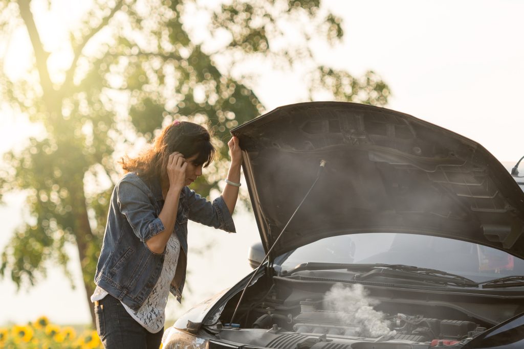 A frustrated woman looking into her car's engine compartment as steam pours out from the overheating engine