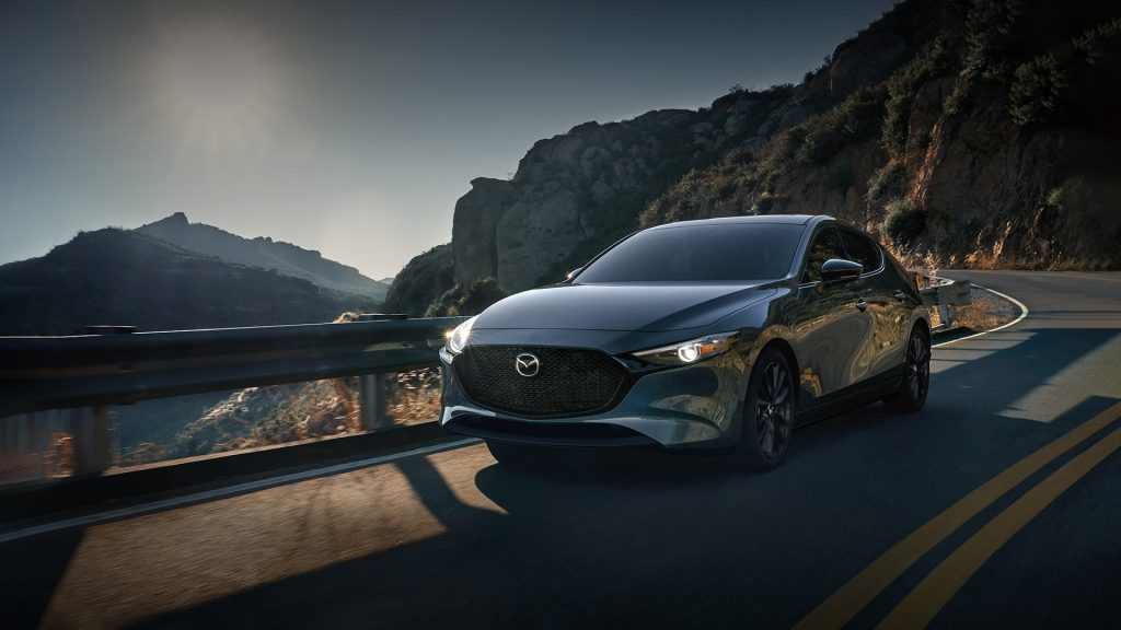 A gray Mazda3 hatchback driving down a mountain road at sunset.