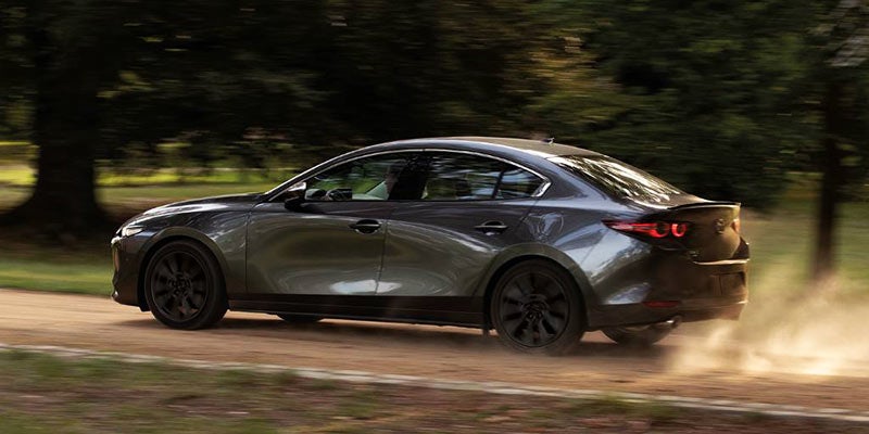 An image of a dark metallic gray Mazda3 sedan rocketing down a country road. 