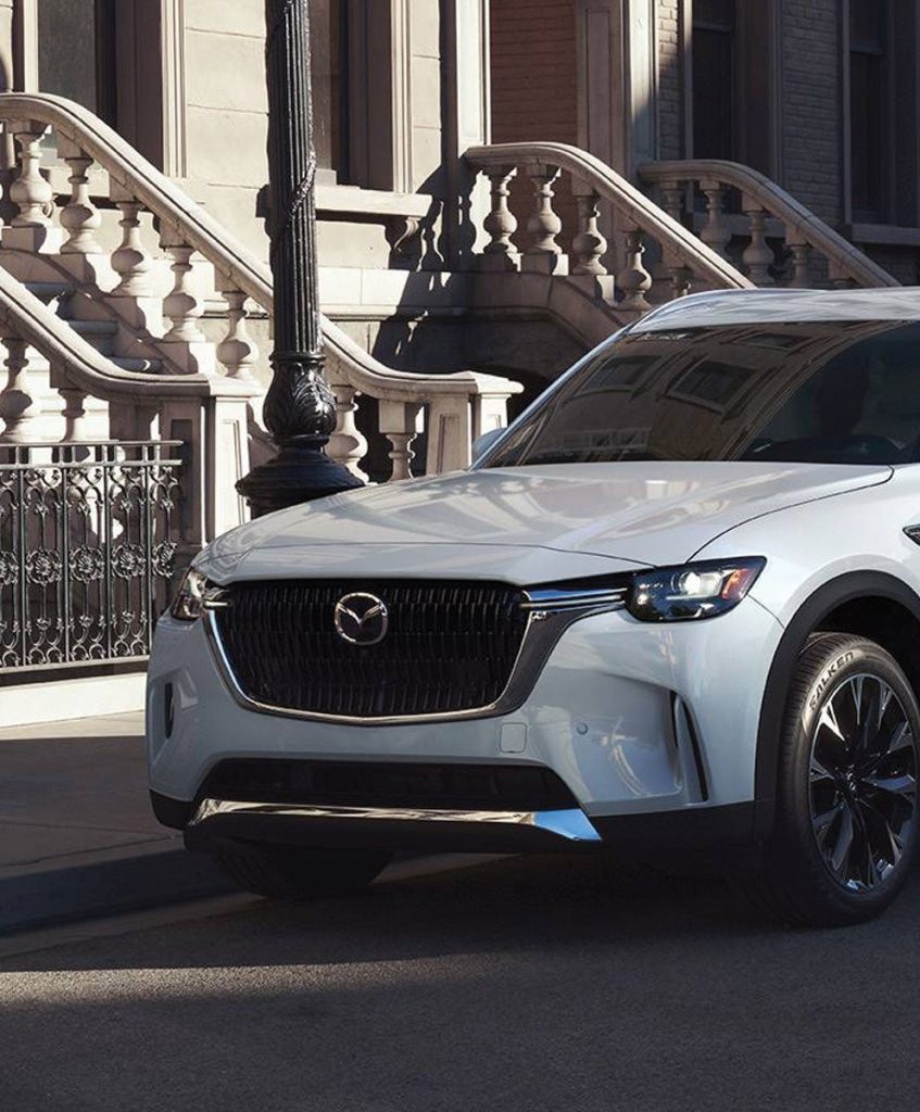 A Mazda CX-90 PHEV parked in front of an apartment building on a city street.