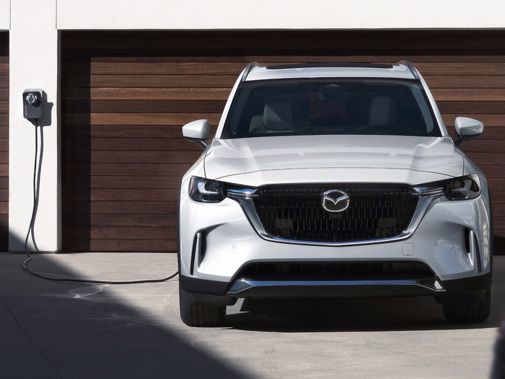 A Mazda CX-90 PHEV sitting in a driveway with a professionally-installed Mazda charger plugged in.