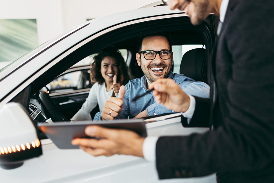 A happy couple seated inside their new vehicle, ready to drive it home from the dealership as a salesperson hands them a clipboard for one last signature.