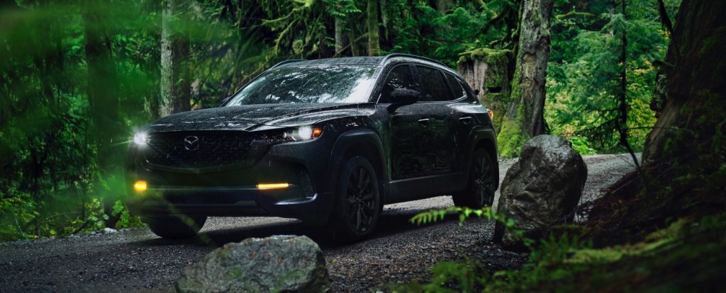 Black Mazda CX-50 with headlights on, driving on a gravel trail through a lush, green forest.